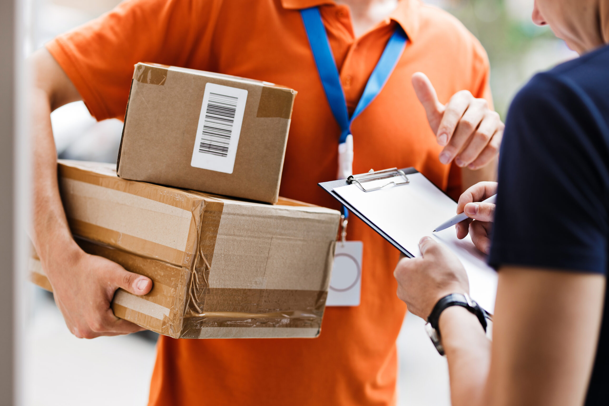 A person wearing an orange T-shirt and a name tag is delivering a parcel to a client, who is putting his signature on the receipt. Friendly worker, high quality delivery service.